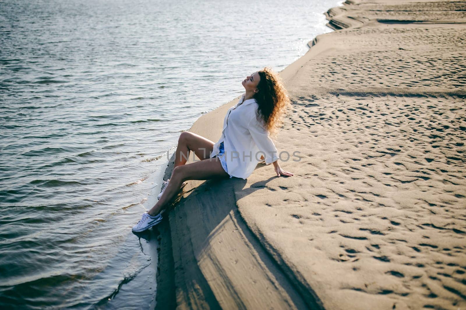 lovely young woman with curly hair, the romance of youth, a journey walk on a warm summer sunny day on a sandy beach with water