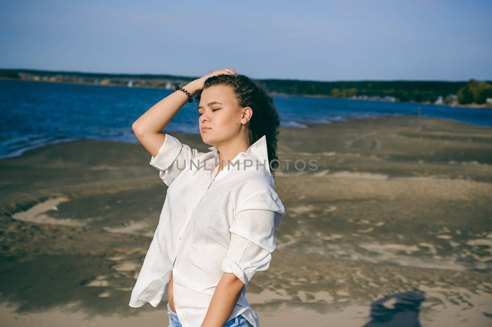 lovely young woman with curly hair, the romance of youth, a journey walk on a warm summer sunny day on a sandy beach with water