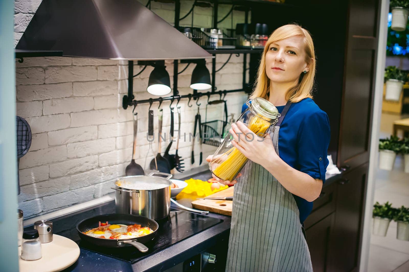 woman housewife preparing in the kitchen. Smiling young blond woman in an apron, holding a glass jar with spaghetti, standing at the stove