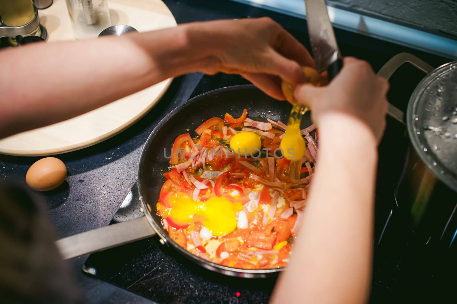 woman housewife prepares fried eggs with ham and vegetables in his kitchen. egg breaks, adding to the pan