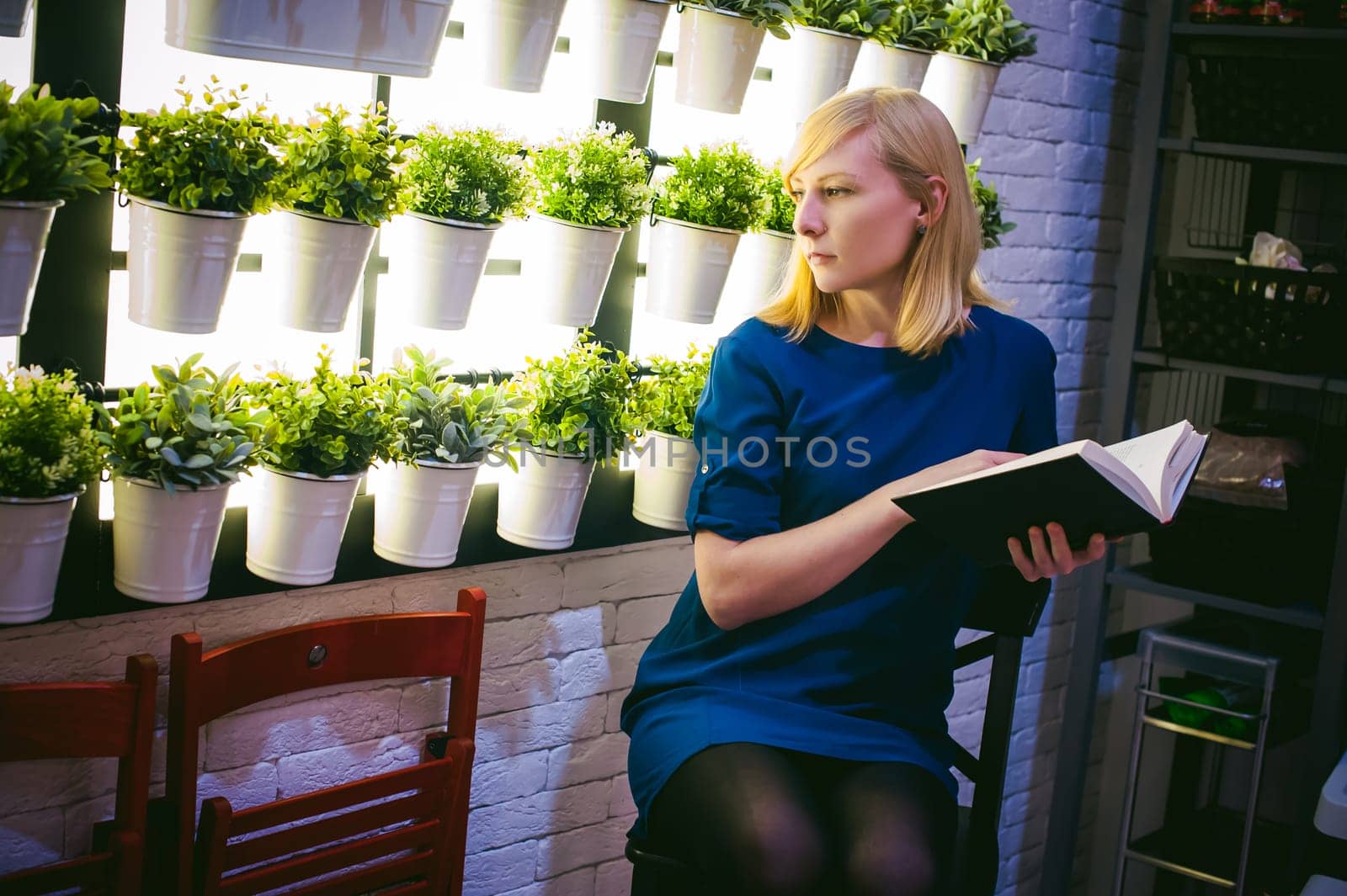 blonde woman in a dress sits on a chair with a book in hand, against the backdrop of houseplants. engaged in reading
