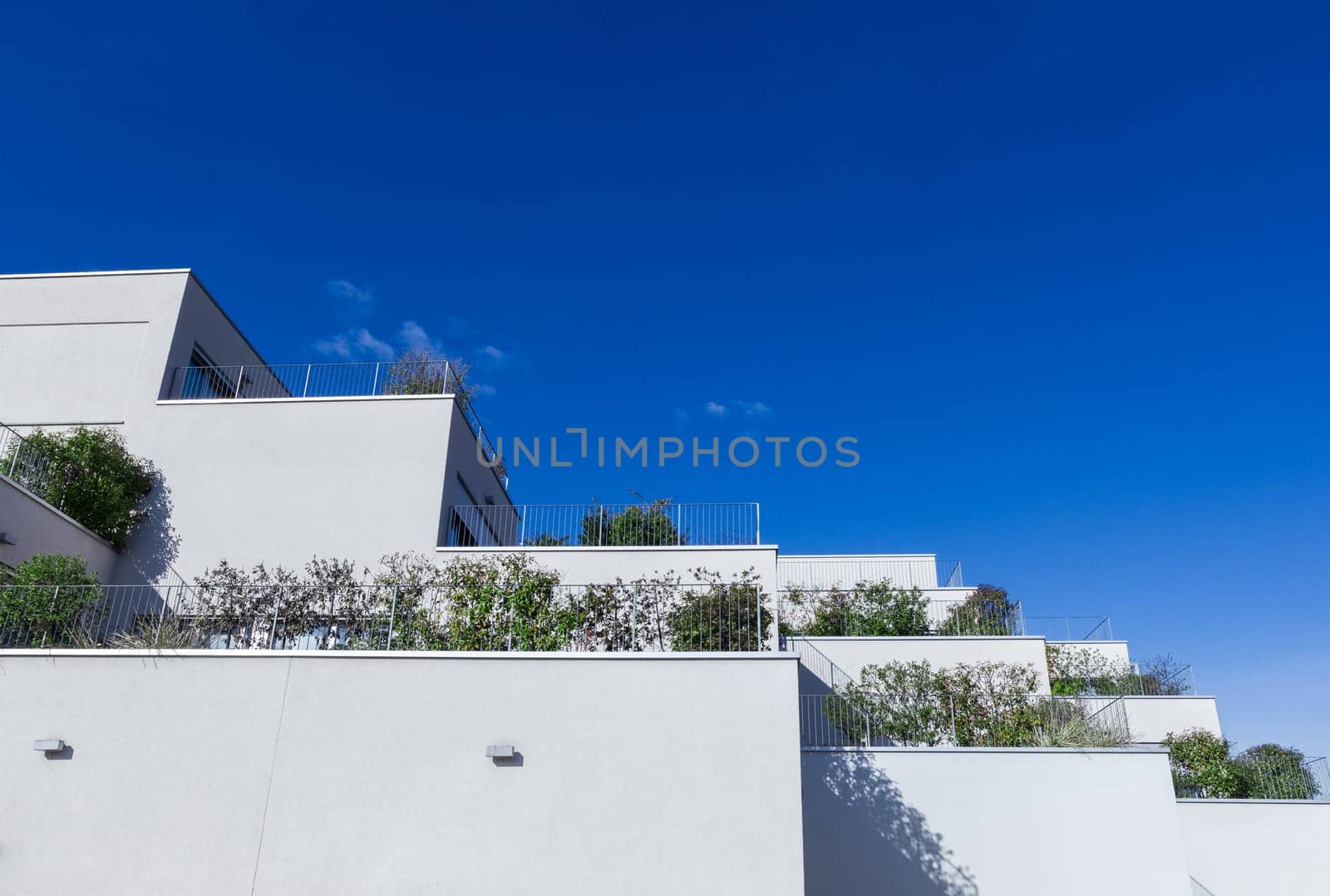 Grey apartment building with modern architecture and large balconies with green plants. Behind a background of blue sky. Bottom view.