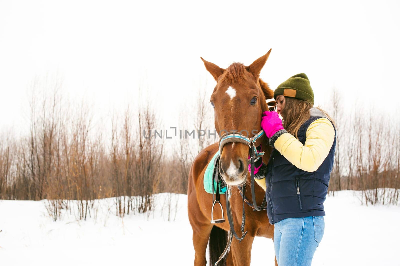 Woman in winter clothes and the horse to stand together against the white snow. love and care for horses. girl puts a bridle on a horse