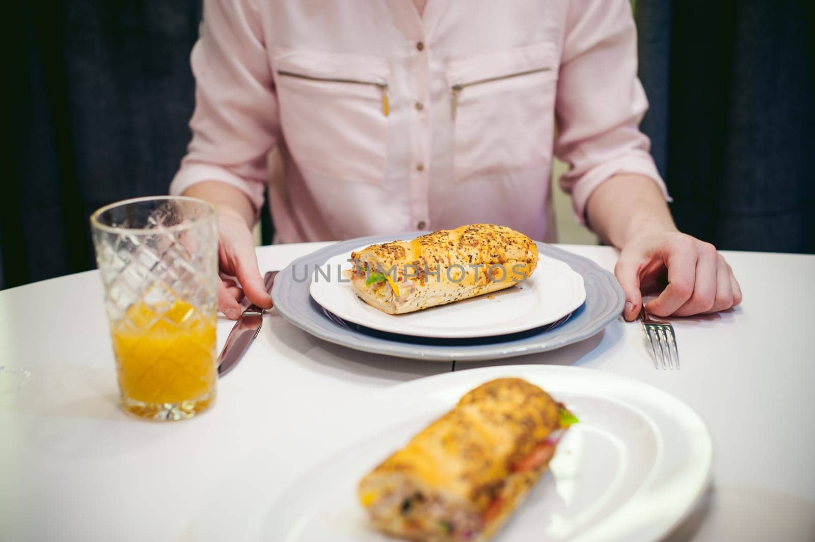 Pretty blonde woman in a pink shirt, eating at home. breakfast sandwich and peach juice. on white table in bright interior of kitchen in plate is sandwich juice poured in glass