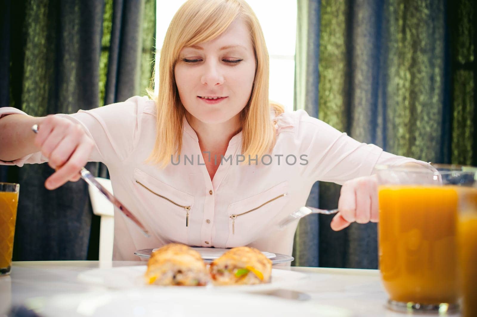 Pretty blonde woman in a pink shirt, eating at home a fork and knife. breakfast sandwich and peach juice. on white table in bright interior of kitchen in plate is sandwich juice poured in glass