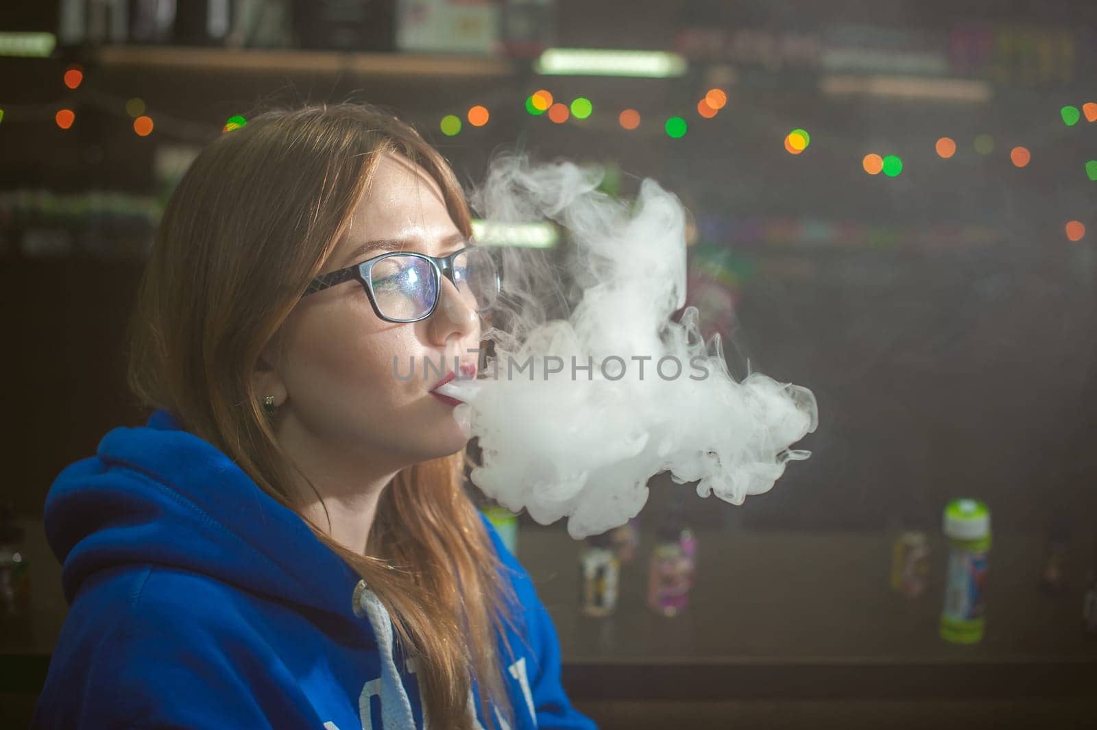 beautiful young woman with glasses smoking electronic cigarette. girl in a blue sweater blows clouds of glycerol vapor, from vape mod, sitting indoors vape shop cafe