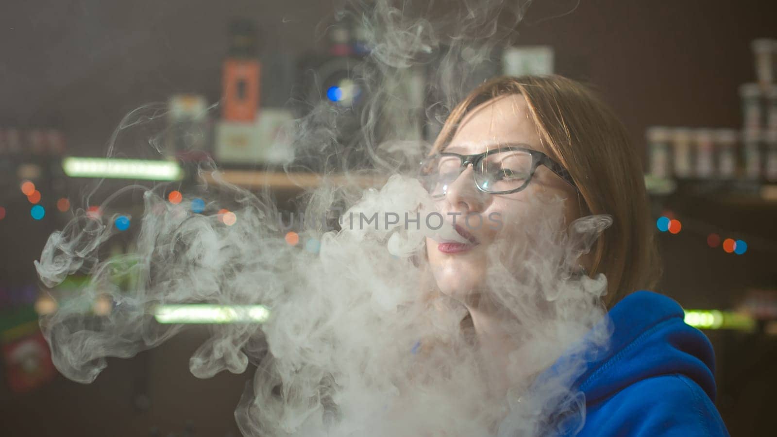 beautiful young woman with glasses smoking electronic cigarette. girl in a blue sweater blows clouds of glycerol vapor, from vape mod, sitting indoors vape shop cafe