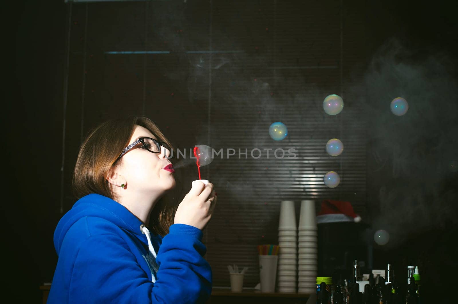young Cute woman, the seller in vape shop shop, smoking electronic cigarette,  vaping and releases a cloud of vapor and inflates soap bubbles