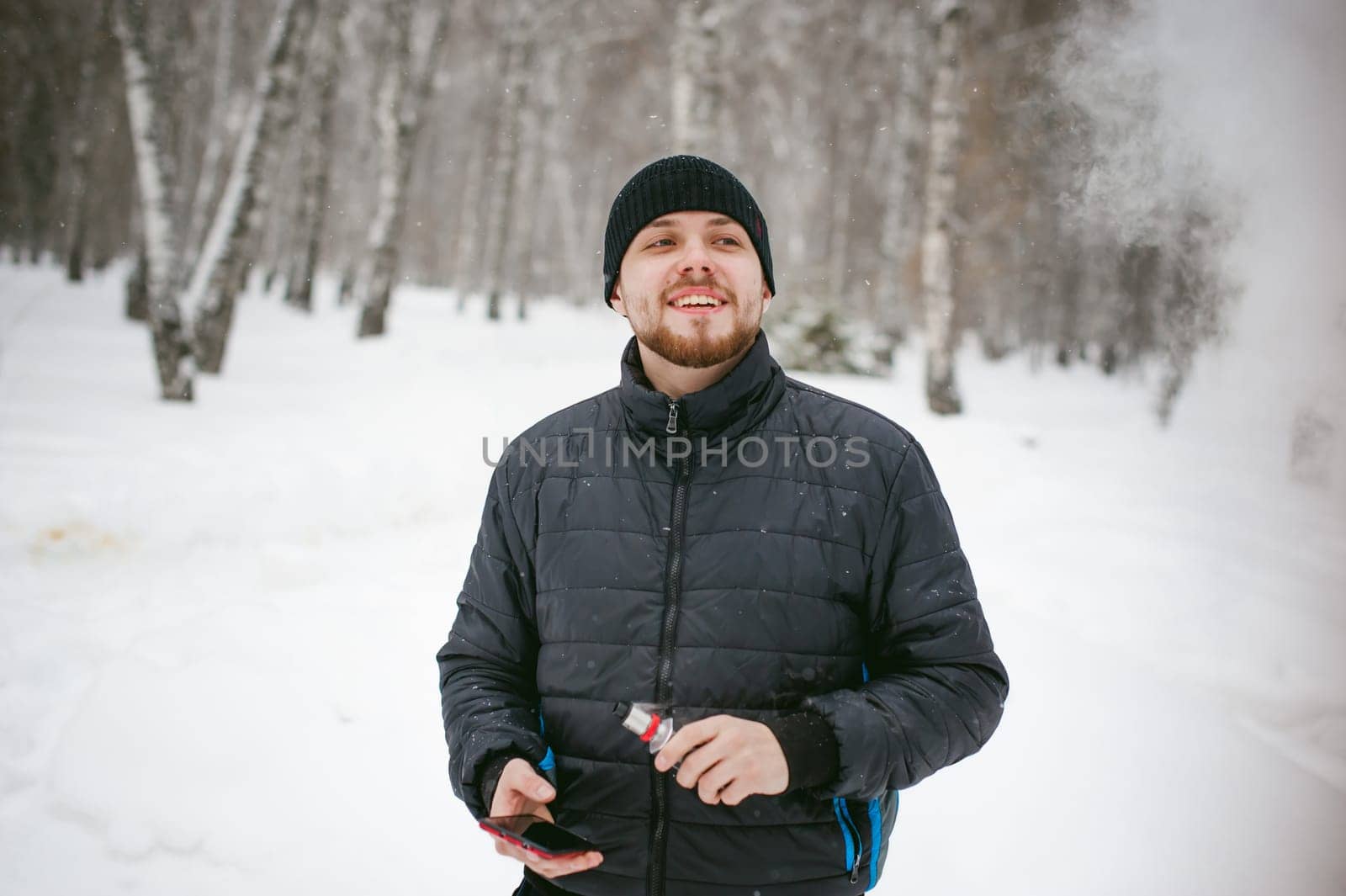 a young man with a beard, smokes an electronic cigarette and happily talking on the phone through the Internet, a positive relaxing outdoors in the snow in the winter