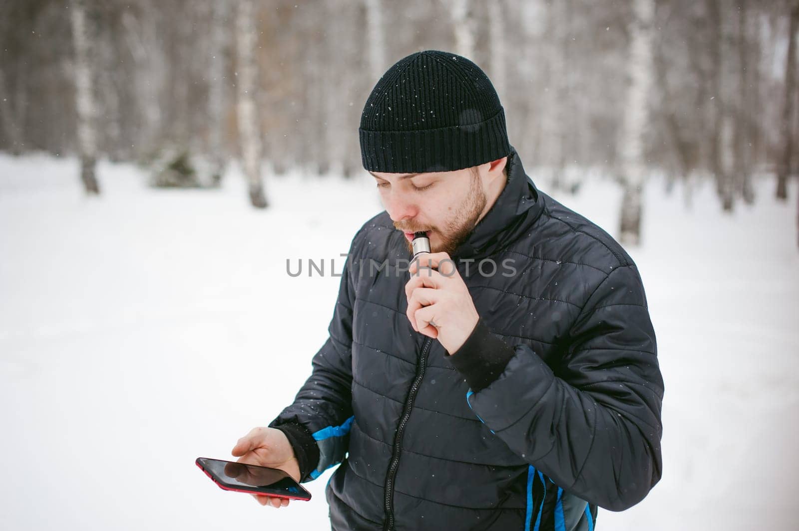 a young man with a beard, smokes an electronic cigarette and happily talking on the phone through the Internet, a positive relaxing outdoors in the snow in the winter