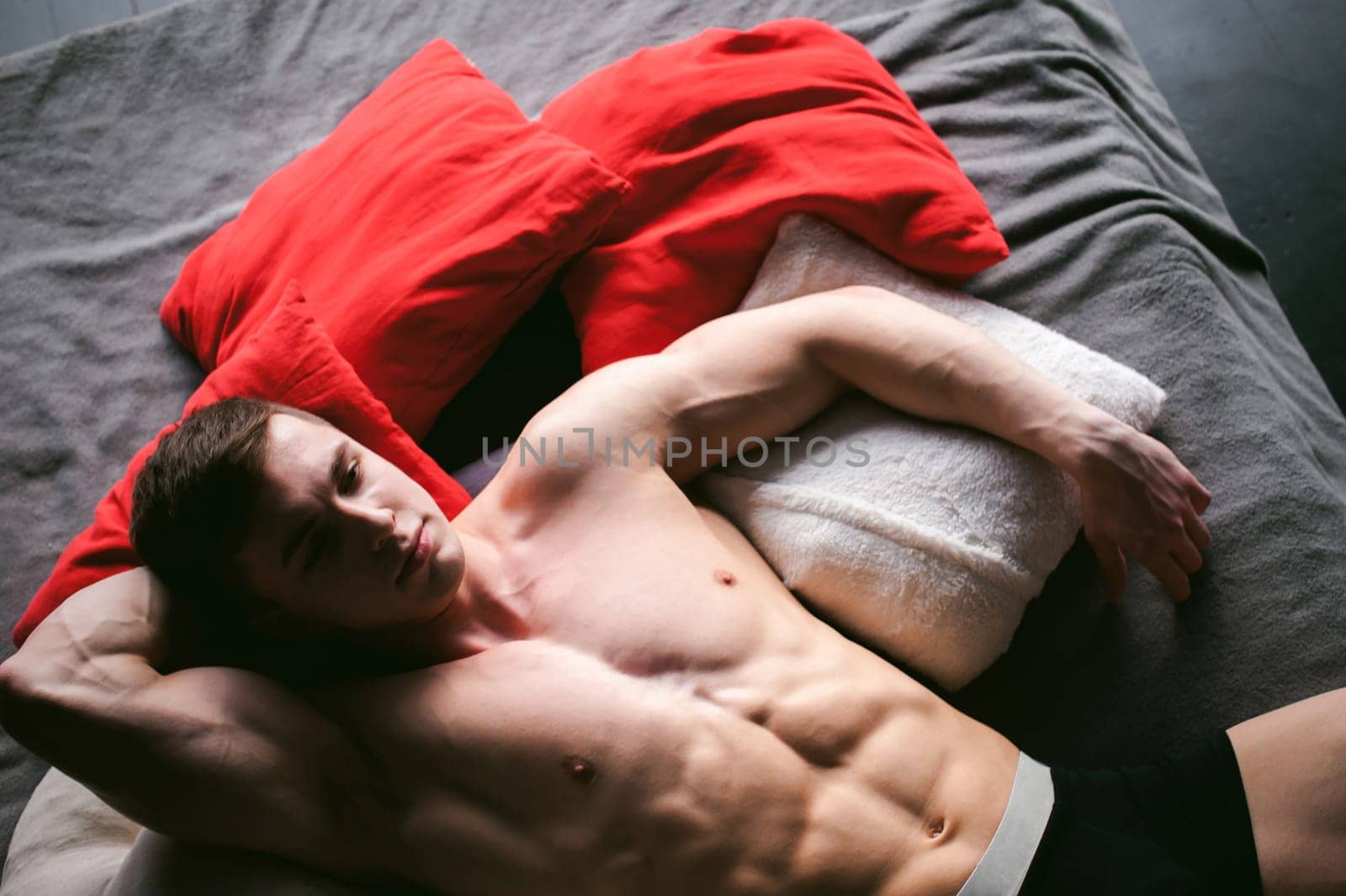 studio portrait young sexy men bodybuilder athlete, with a bare torso, lies on a bed in pillows in underwear