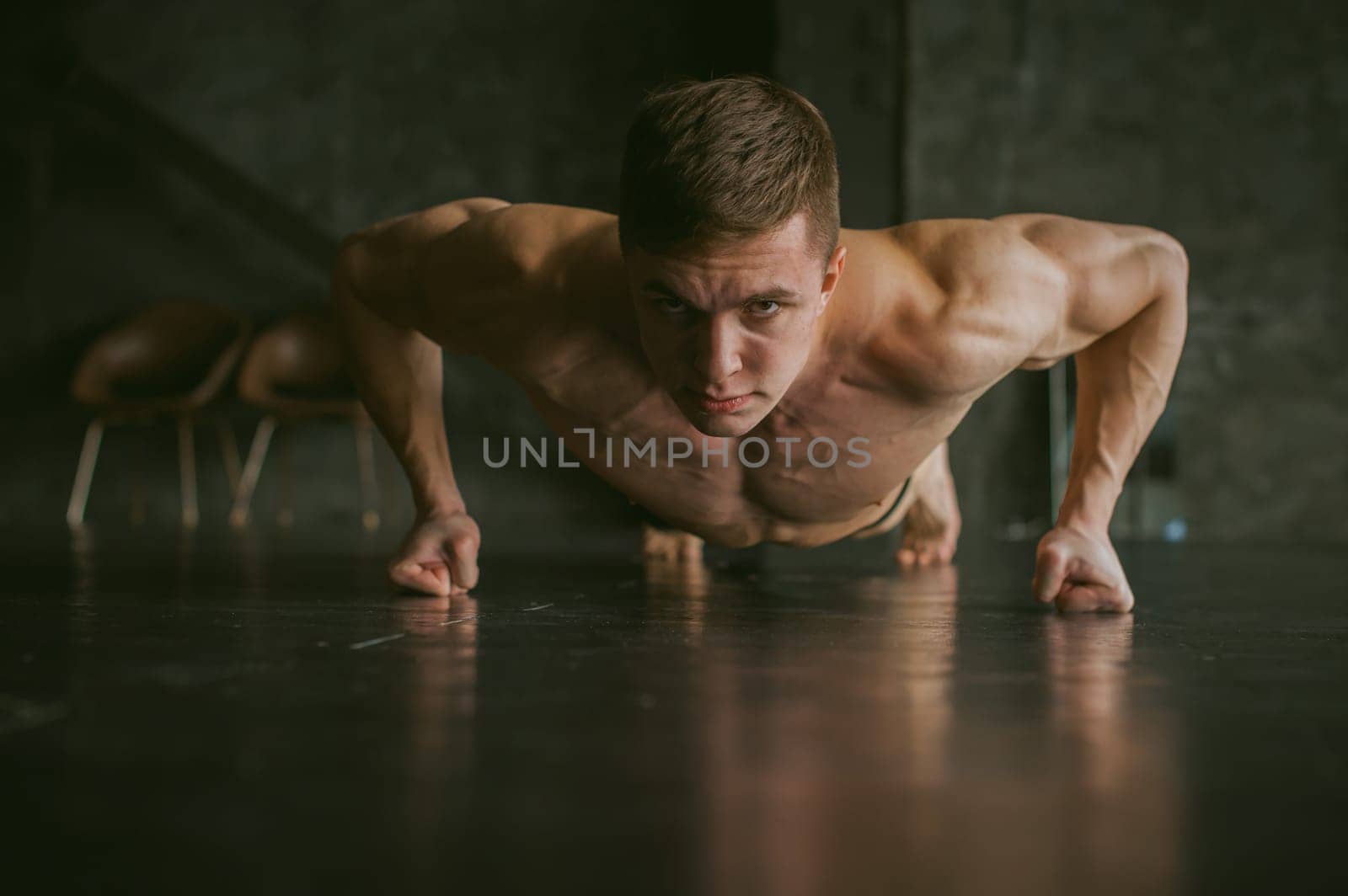 Young sexy male bodybuilder athlete with a bare torso. portrait of a studio of a light-skinned strong man engaged in push-ups on the fists, on the floor, straining powerful hands