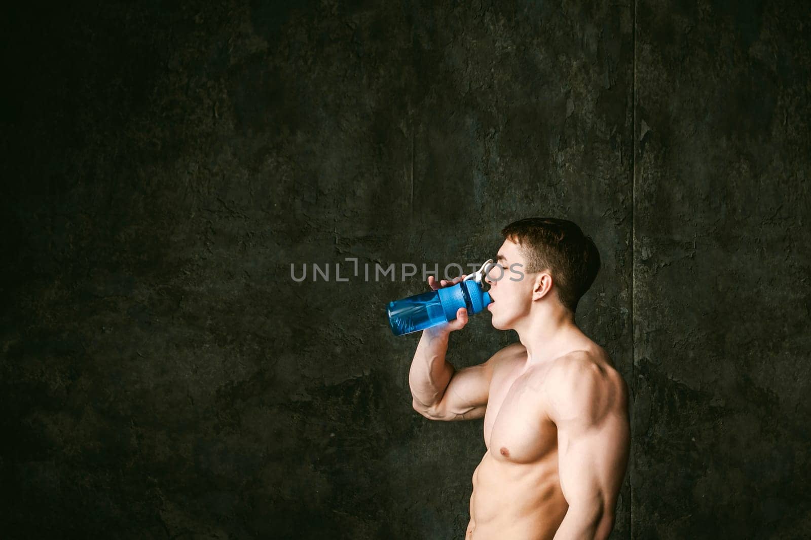 Young sexy men bodybuilder athlete,studio portrait loft on background of stylized wall,guy model With a bare torso Drinks water from a sports bottle of blue color after training