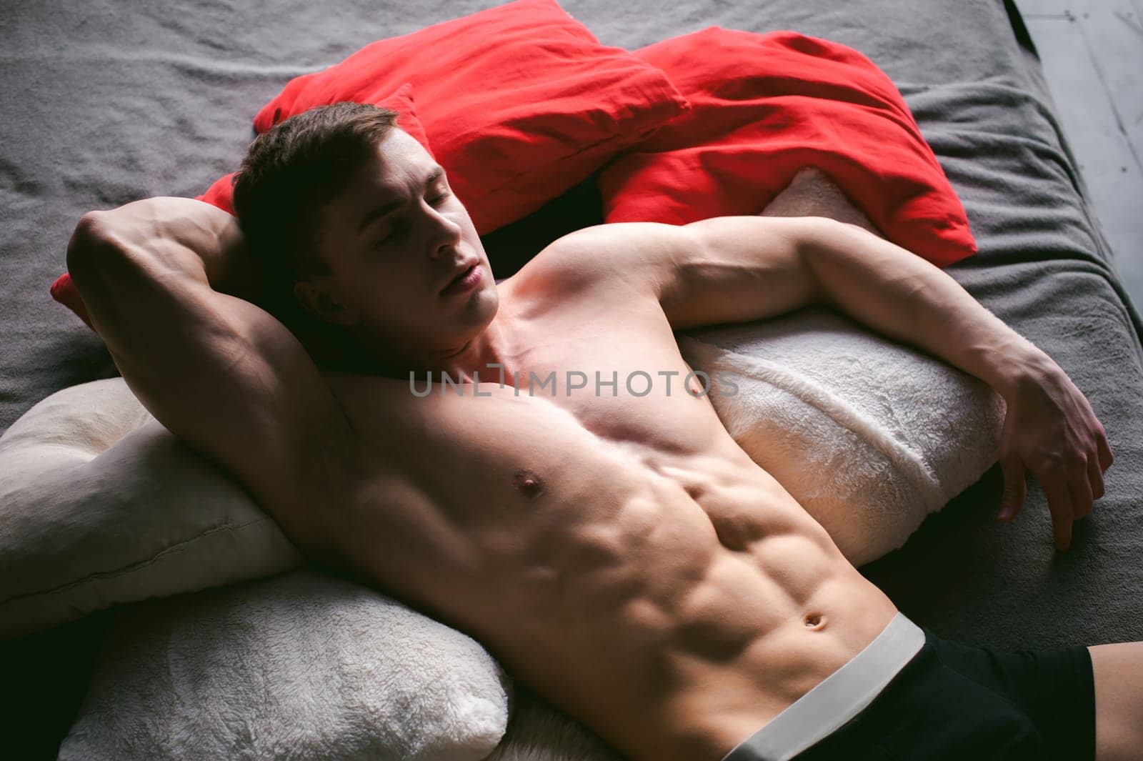 studio portrait young sexy men bodybuilder athlete, with a bare torso, lies on a bed in pillows in underwear
