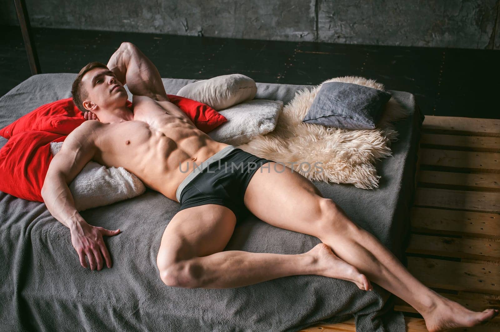 studio portrait young sexy men bodybuilder athlete, with a bare torso, lies on a bed in pillows in underwear