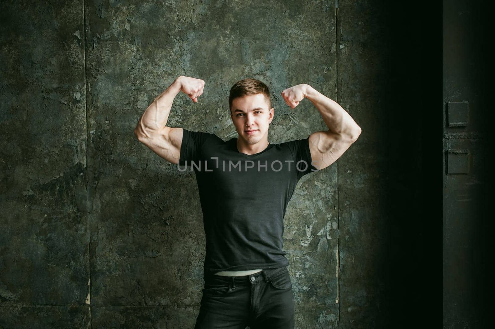 Young sexy men bodybuilder athlete, studio portrait loft on background of stylized Concrete brutal wall, guy model black Tshirt and trousers With powerful muscular arms demonstrates strength