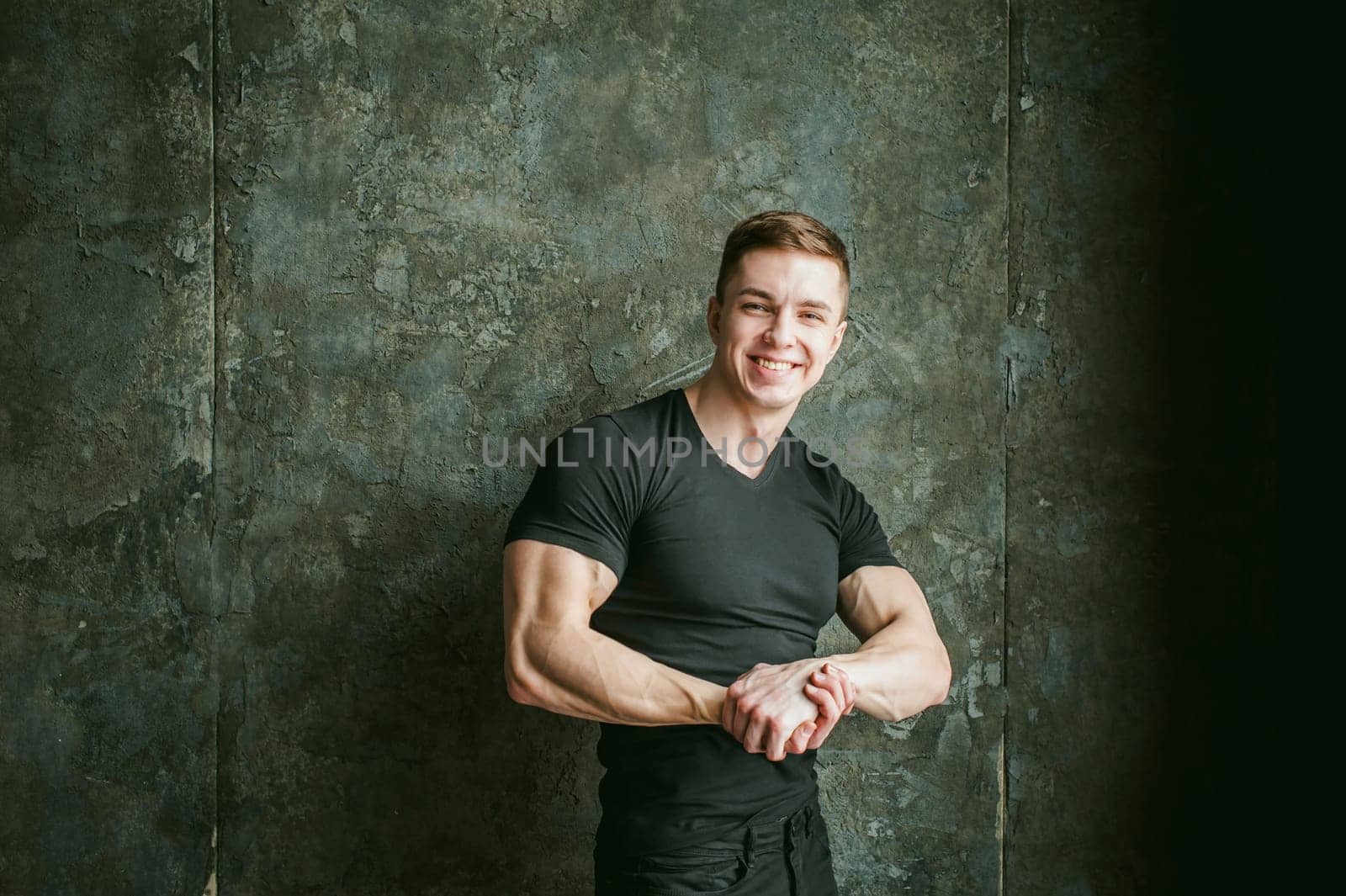 Young sexy men bodybuilder athlete, studio portrait loft on background of stylized Concrete brutal wall, guy model black Tshirt and trousers With powerful muscular arms demonstrates strength