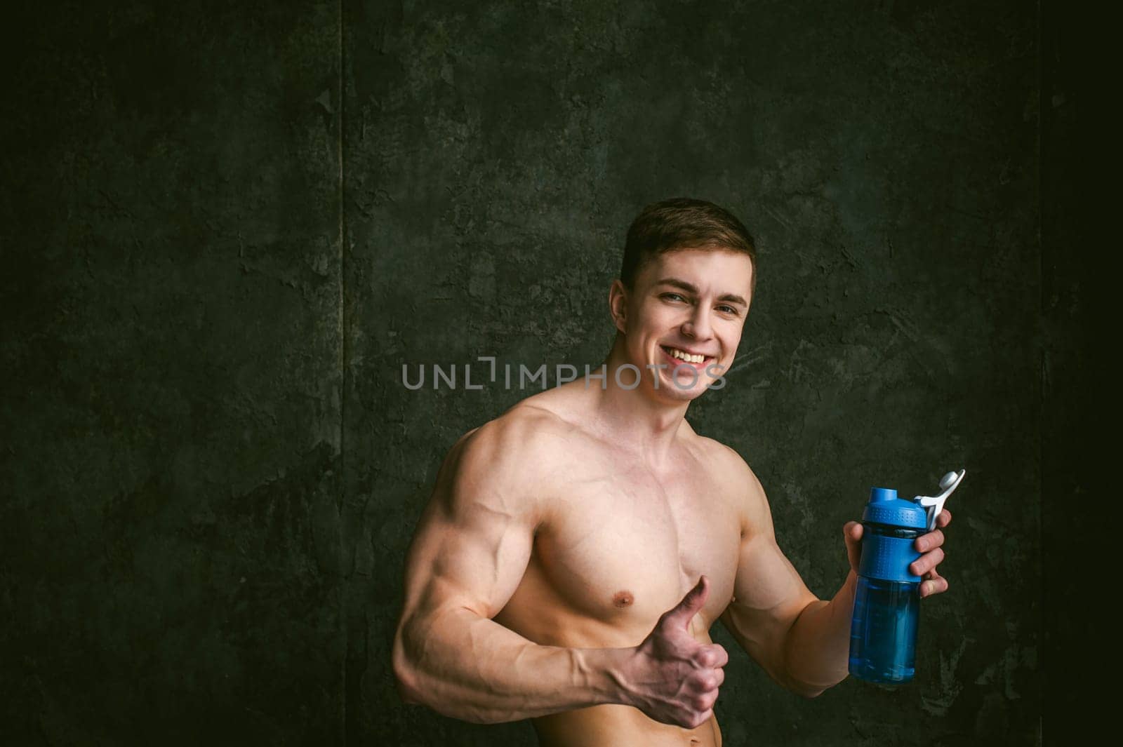 Young sexy men bodybuilder athlete,portrait loft on background of stylized wall,guy model With bare torso Holds in his hand blue sports bottle for drinking during training, show thumbs up and smiling