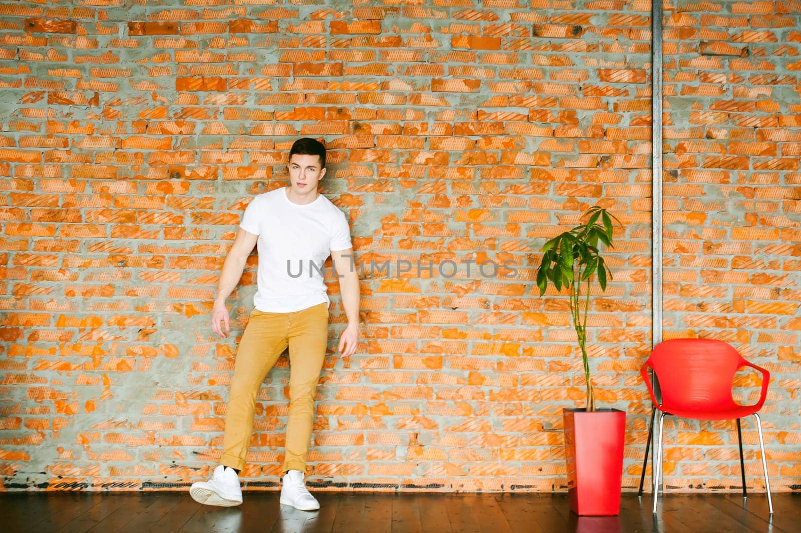 Young sexy men bodybuilder athlete,studio portrait in loft, guy model in white T-shirt and brown trousers, Leaned against a brick wall