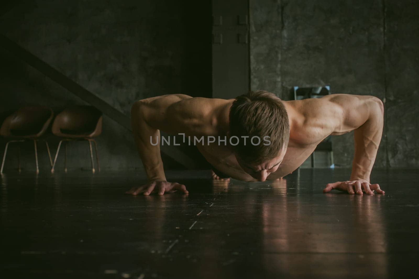 Young sexy male bodybuilder athlete with a bare torso. portrait of a studio of a light-skinned strong man engaged in push-ups, on the floor, straining powerful hands