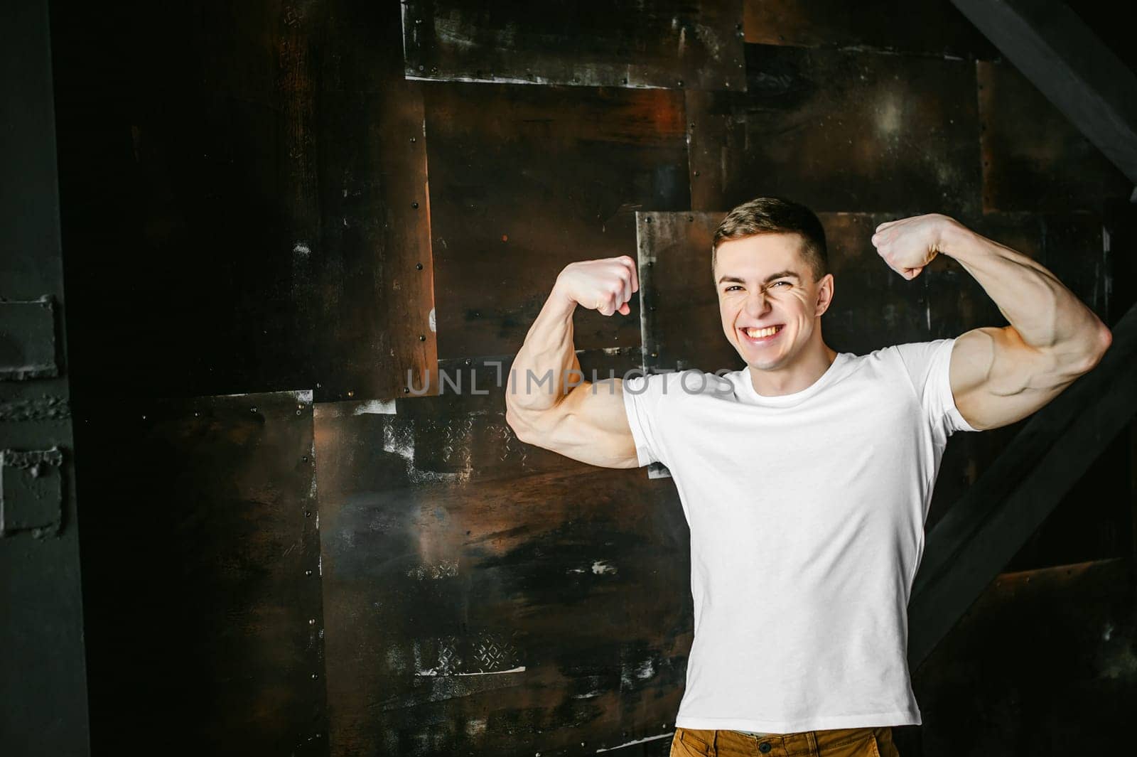 Young sexy men bodybuilder athlete, studio portrait in a loft on a background of  stylized metal wall, a guy model in a white T-shirt and brown trousers, tensed his arms showing muscles, bared teeth