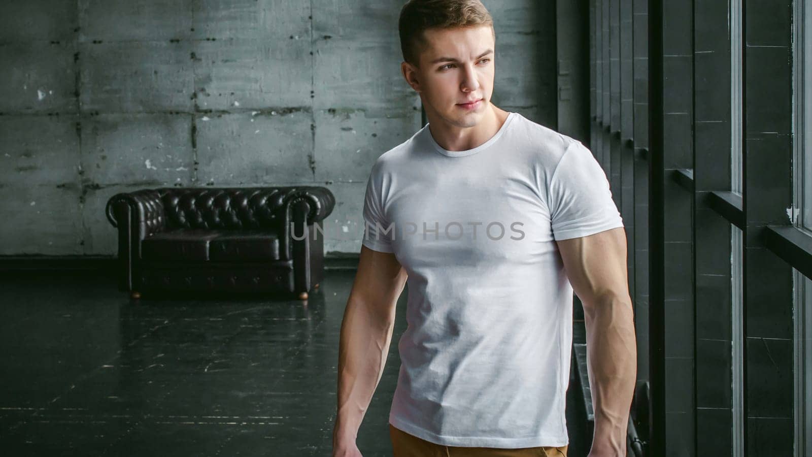 Young sexy men bodybuilder athlete,studio portrait in loft on the background of stylized wall and Black leather sofa, guy model in white T-shirt and brown trousers against background of larger window