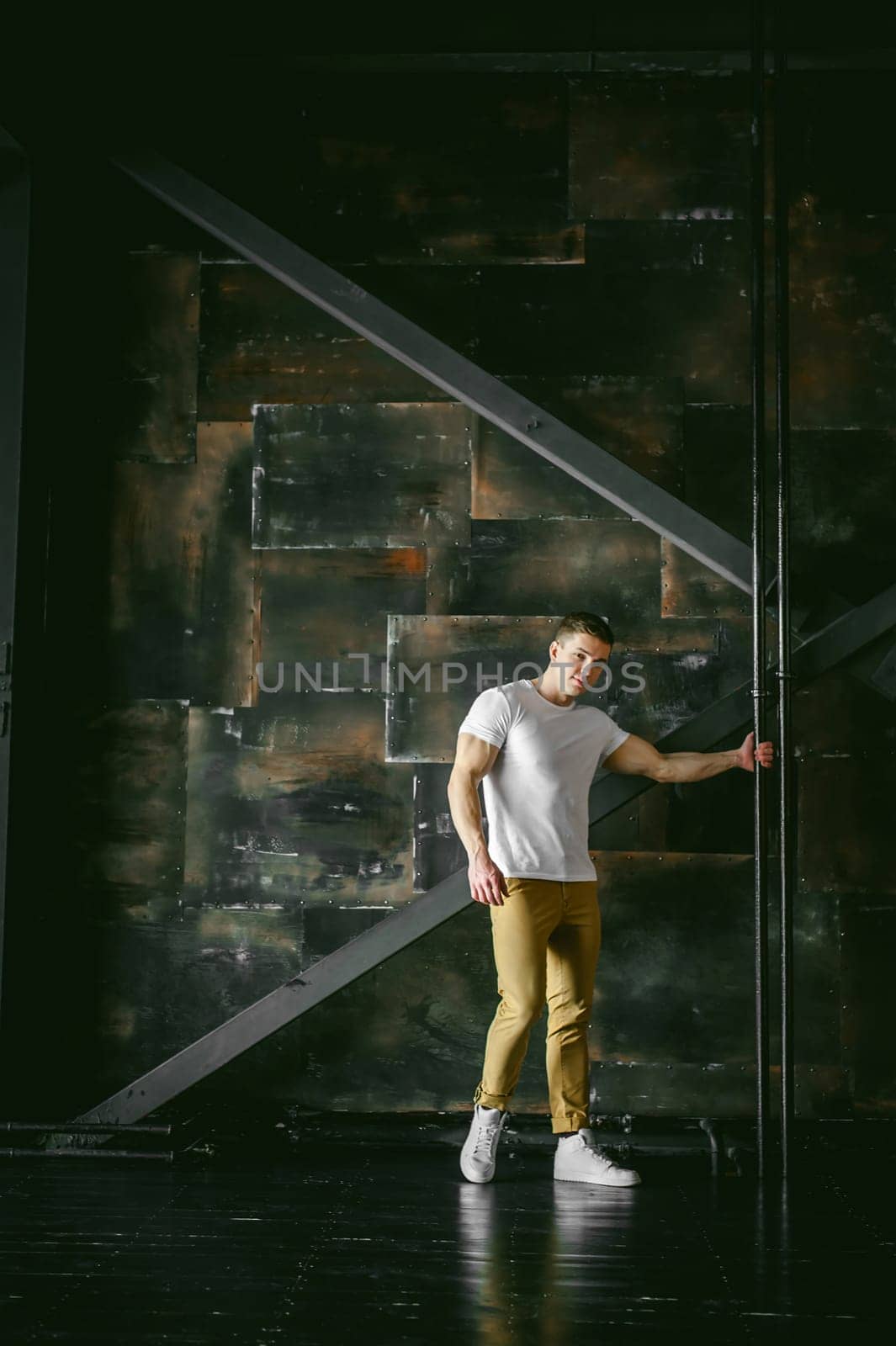 Young sexy men bodybuilder athlete, Studio portrait in a loft on the background of a stylized metal wall, a guy model in a white T-shirt and brown trousers