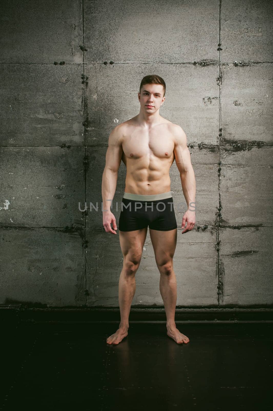 Young sexy male bodybuilder sportsman with a bare torso against a gray wall background, studio portrait of a light-skinned strong man in full growth