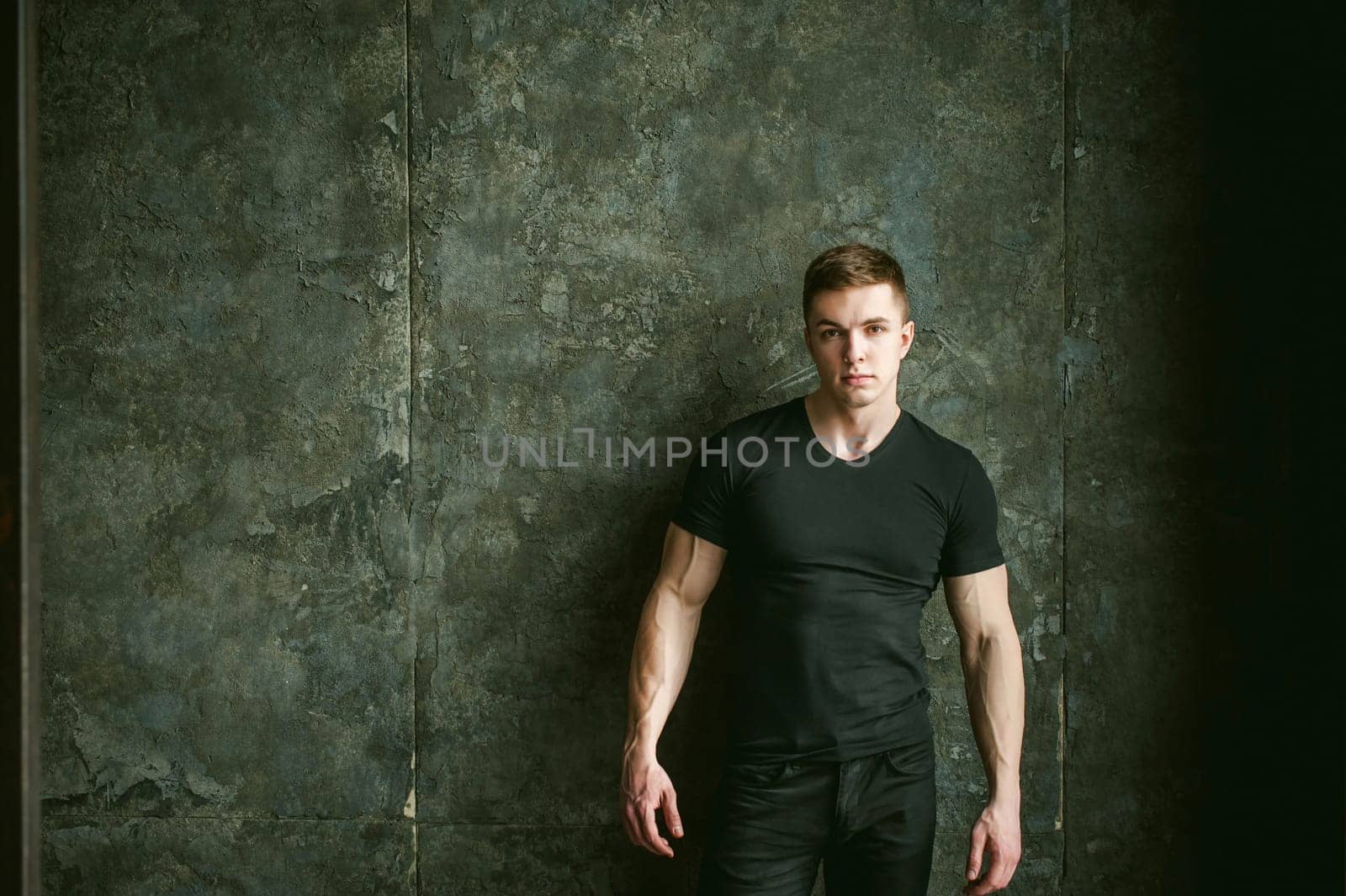 Young sexy men bodybuilder athlete, studio portrait loft on background of stylized Concrete brutal wall, guy model black Tshirt and trousers With powerful muscular arms