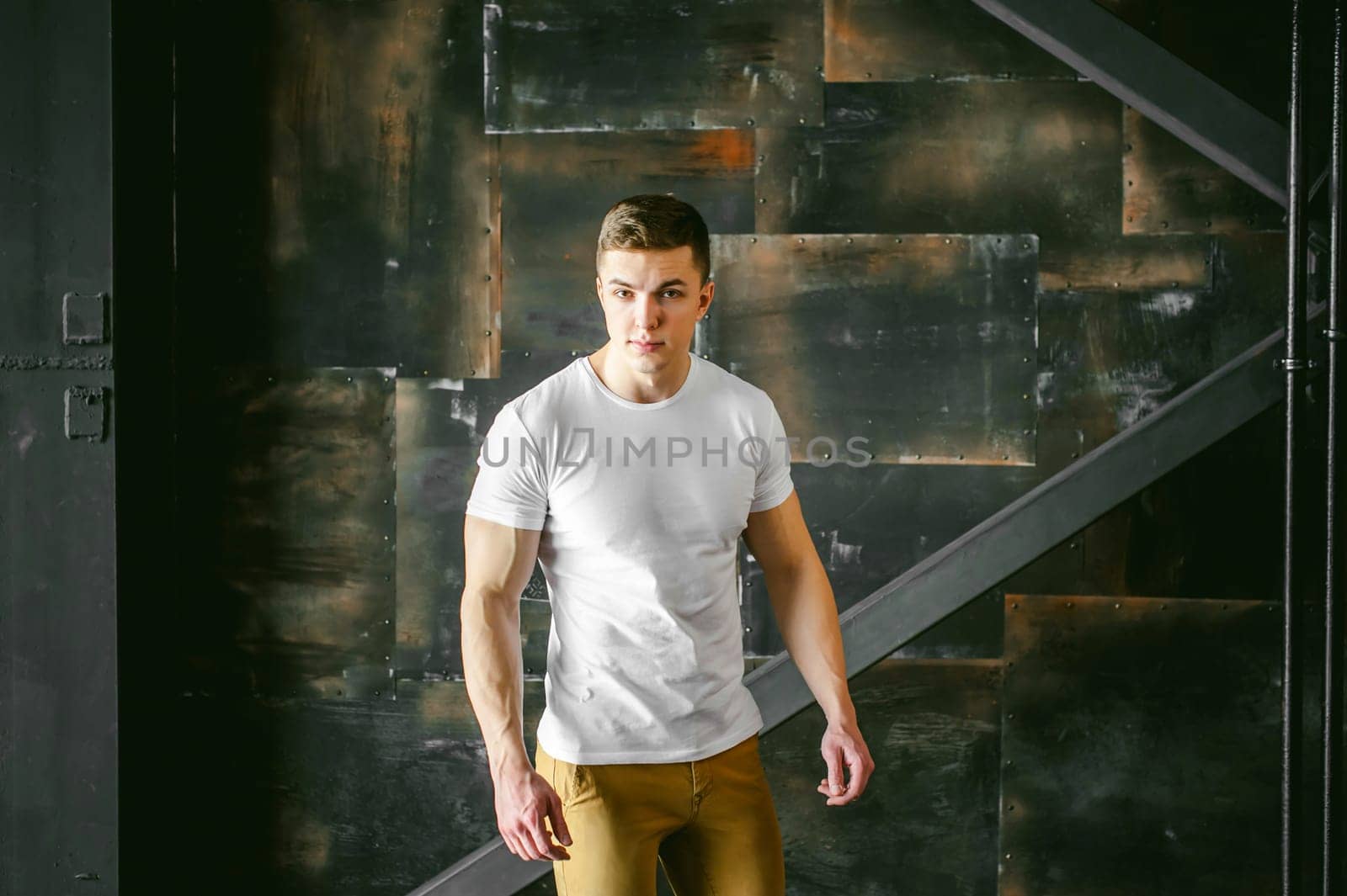 Young sexy men bodybuilder athlete, Studio portrait in a loft on the background of a stylized metal wall, a guy model in a white T-shirt and brown trousers