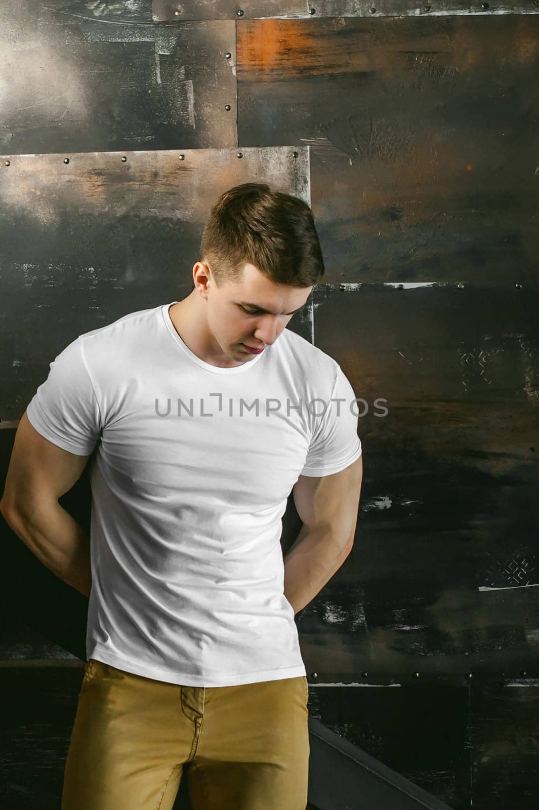 Young sexy men bodybuilder athlete, Studio portrait in a loft on the background of a stylized metal wall, a guy model in a white T-shirt and brown trousers
