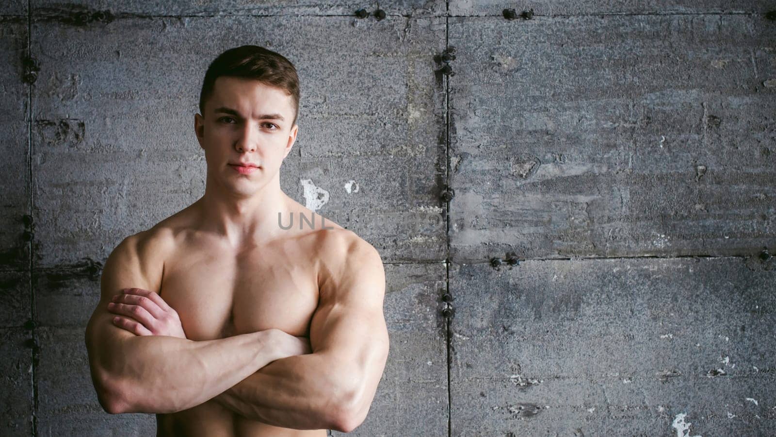 Young sexy male bodybuilder athlete with a bare torso against a gray brutal concrete wall, portrait of a studio of a light-skinned strong man crossed powerful arms on his chest