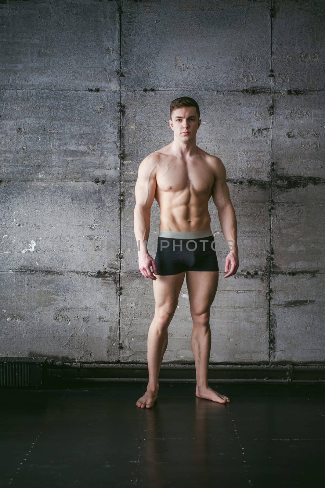 Young sexy male bodybuilder sportsman with a bare torso against a gray wall background, studio portrait of a light-skinned strong man in full growth