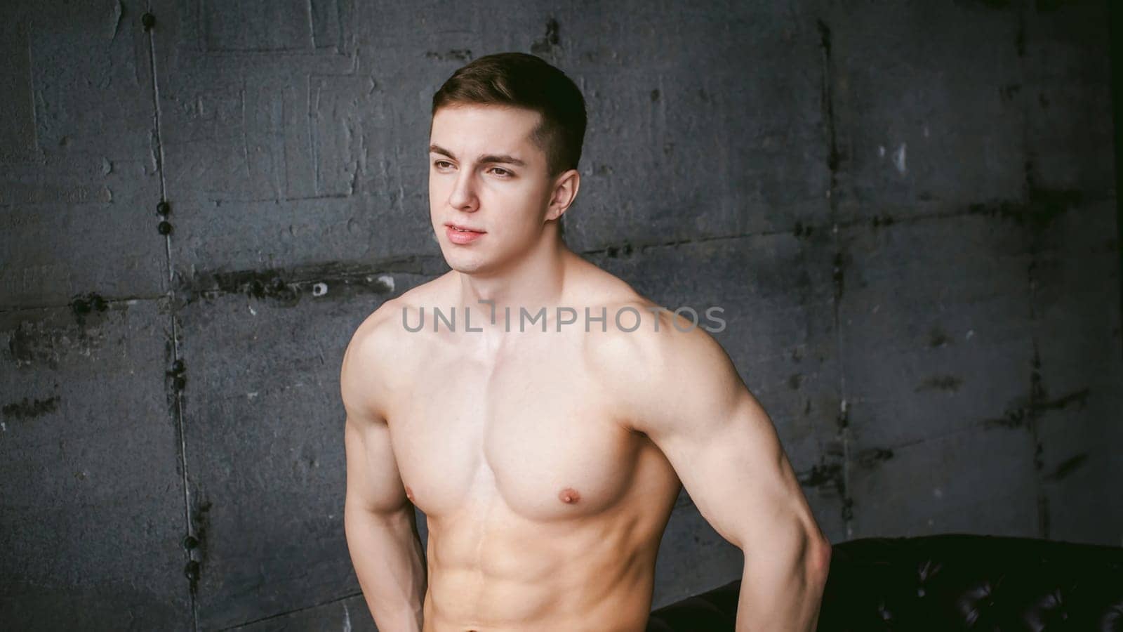 Young sexy male bodybuilder athlete with bare torso against a gray brutal concrete wall, studio portrait of a light-skinned strong man