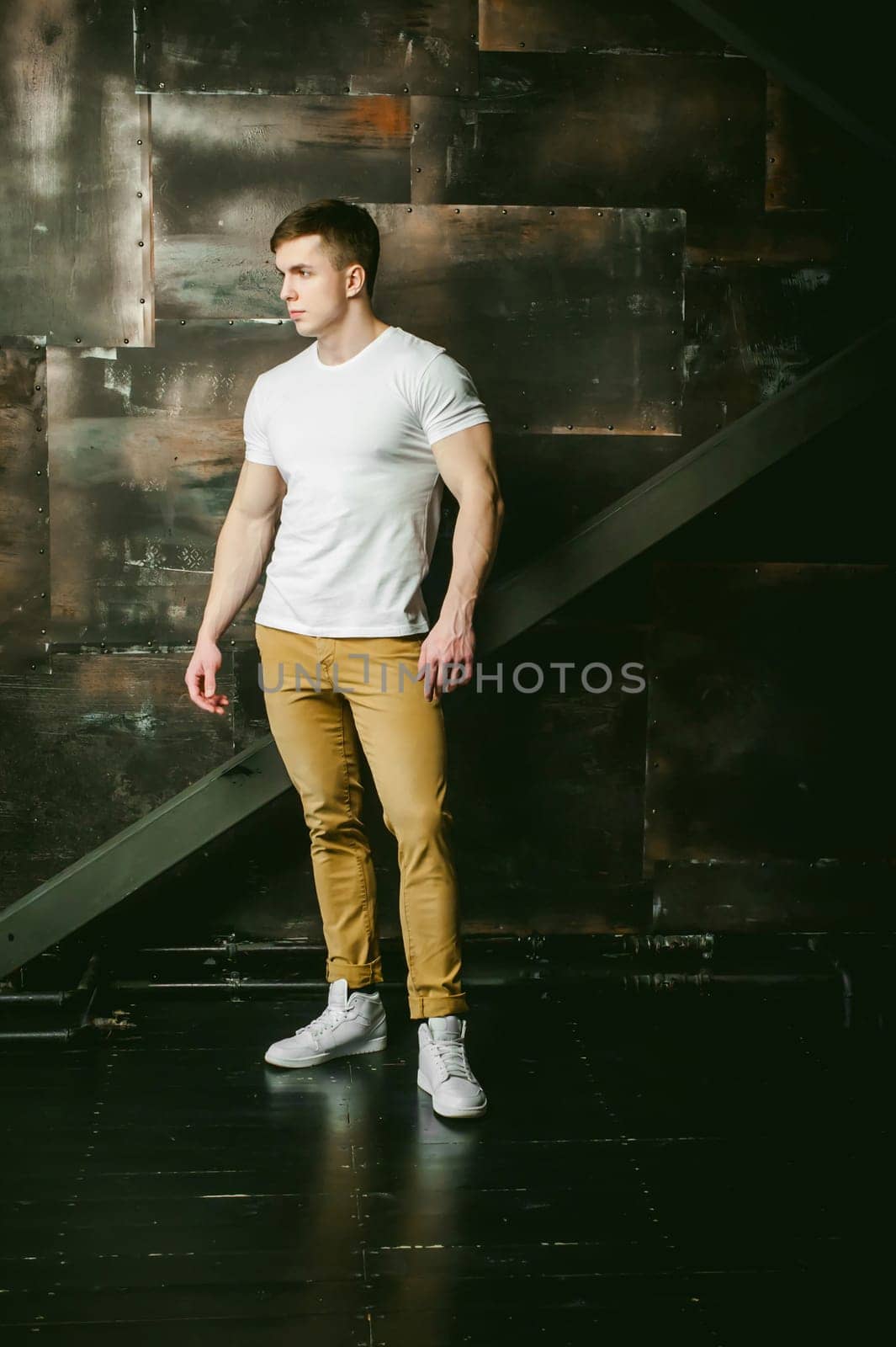 Young sexy men bodybuilder athlete, Studio portrait in a loft on the background of a stylized metal wall, a guy model in a white T-shirt and brown trousers