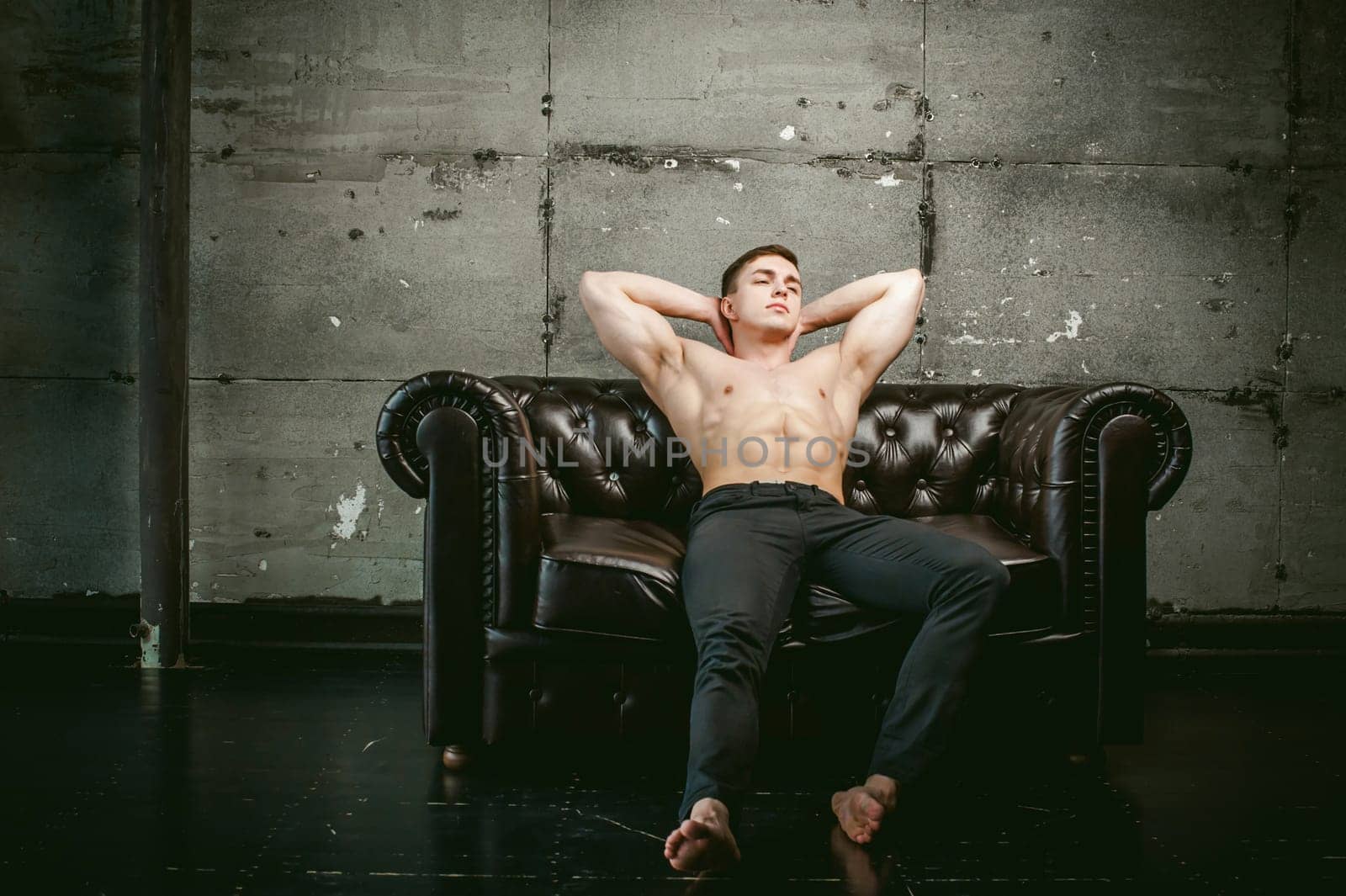 Young sexy men bodybuilder athlete sitting on a black leather couch with a naked torso and black trousers, Threw one's hands behind his head, studio portrait