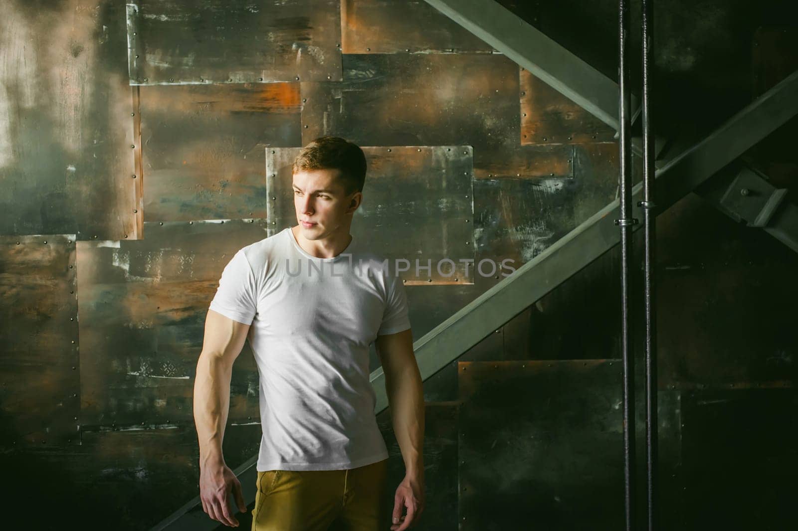 Young sexy men bodybuilder athlete, Studio portrait in a loft on the background of a stylized metal wall, a guy model in a white T-shirt and brown trousers