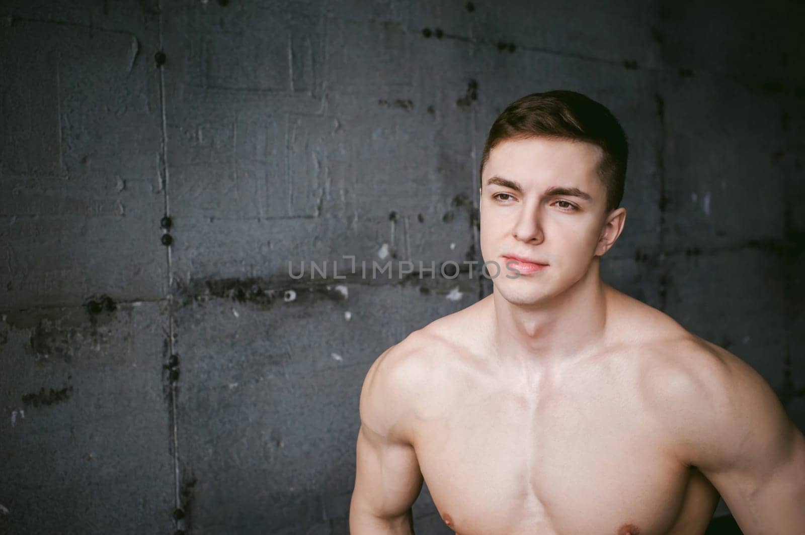 Young sexy male bodybuilder athlete with bare torso against a gray brutal concrete wall, studio portrait of a light-skinned strong man