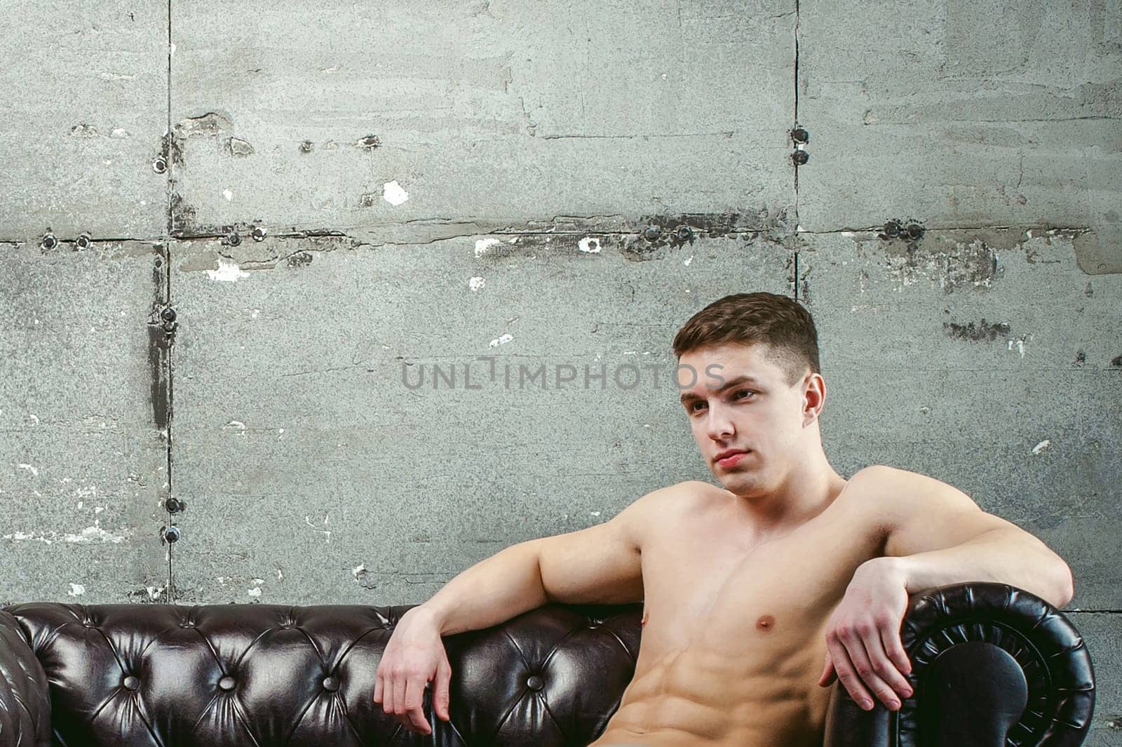 Young sexy men bodybuilder athlete sitting on a black leather couch with a naked torso, studio portrait