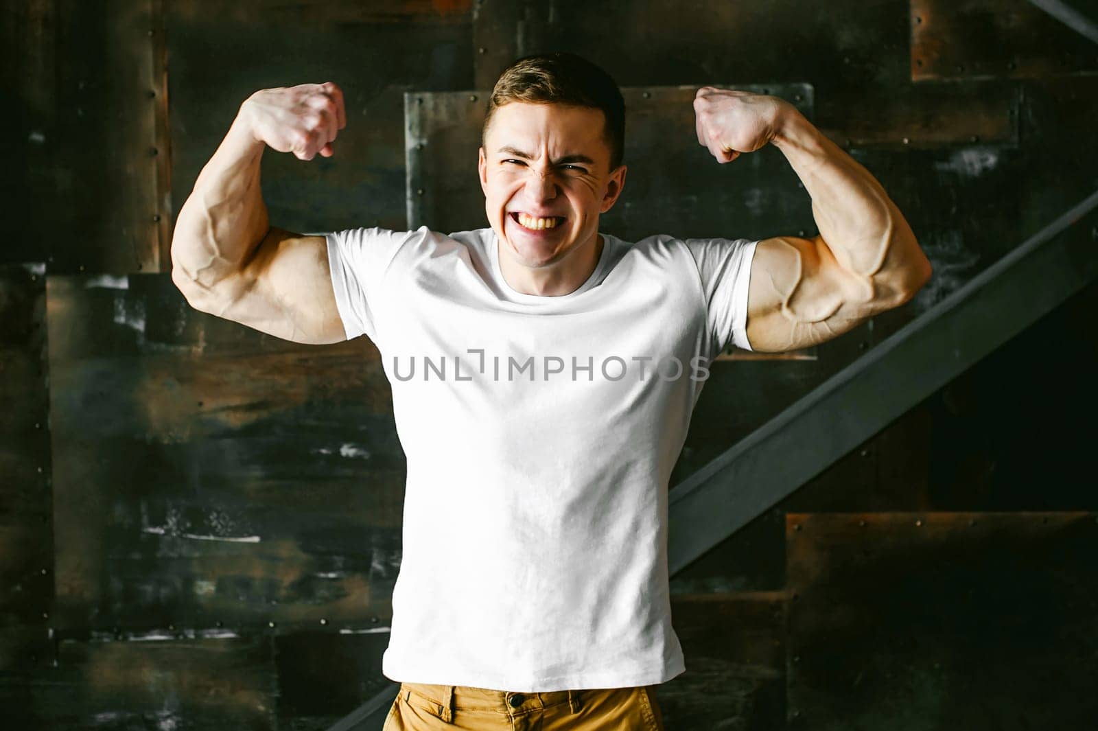 Young sexy men bodybuilder athlete, studio portrait in a loft on a background of  stylized metal wall, a guy model in a white T-shirt and brown trousers, tensed his arms showing muscles, bared teeth