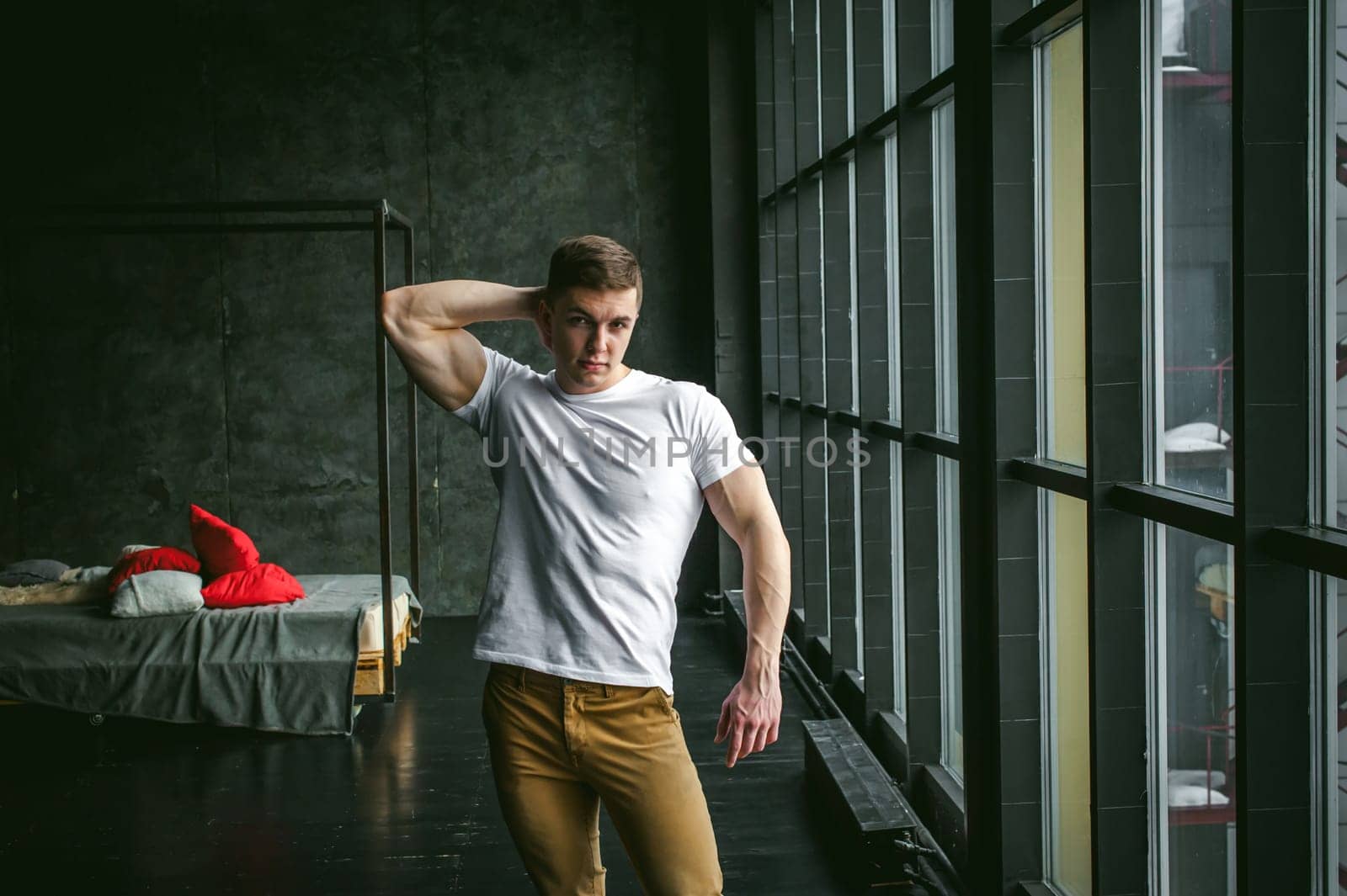 Young sexy men bodybuilder athlete, studio portrait in loft on the background of stylized wall and brutal bed, guy model in white T-shirt and brown trousers against the background of a larger window