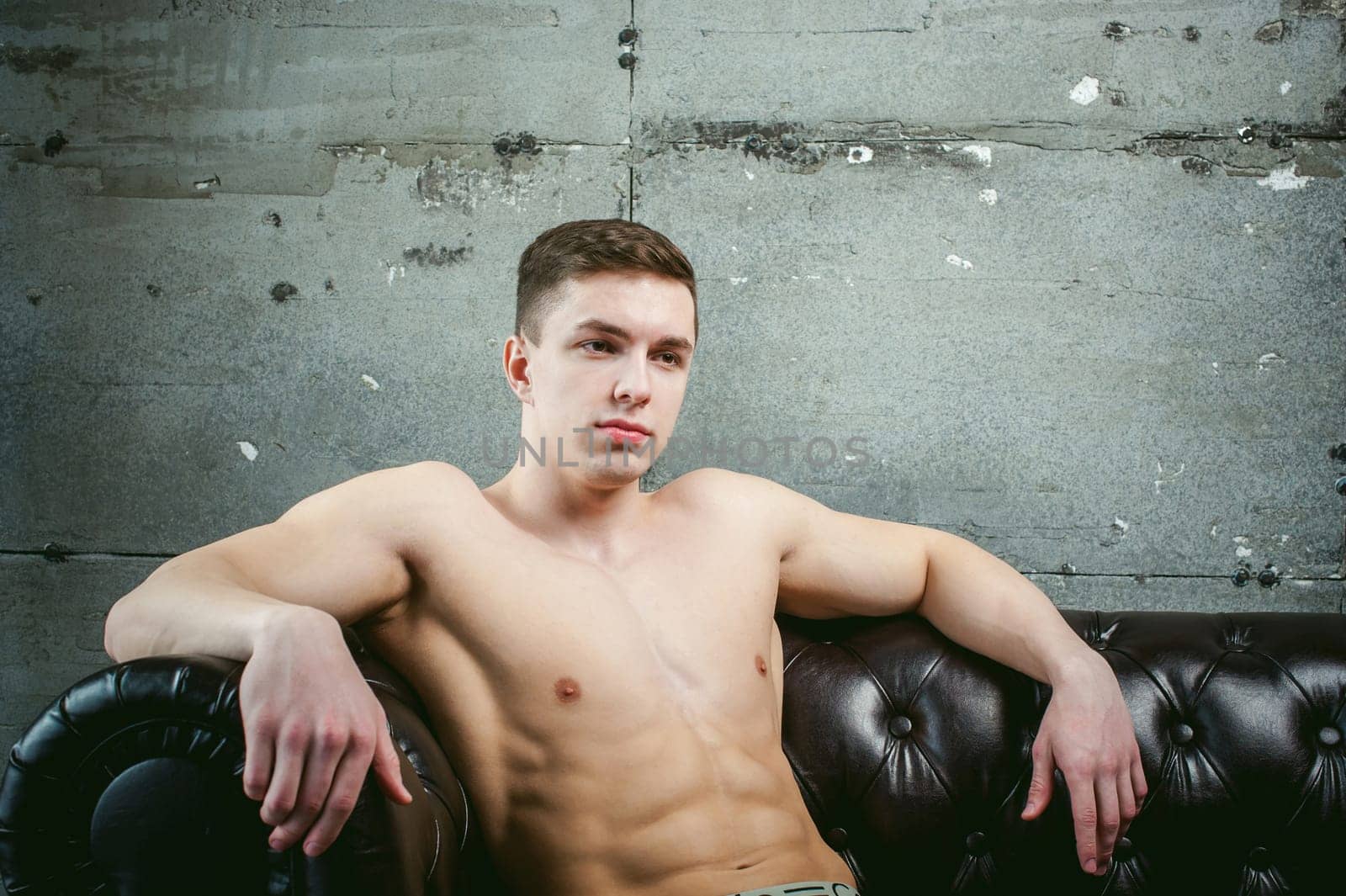 Young sexy men bodybuilder athlete sitting on a black leather couch with a naked torso, studio portrait