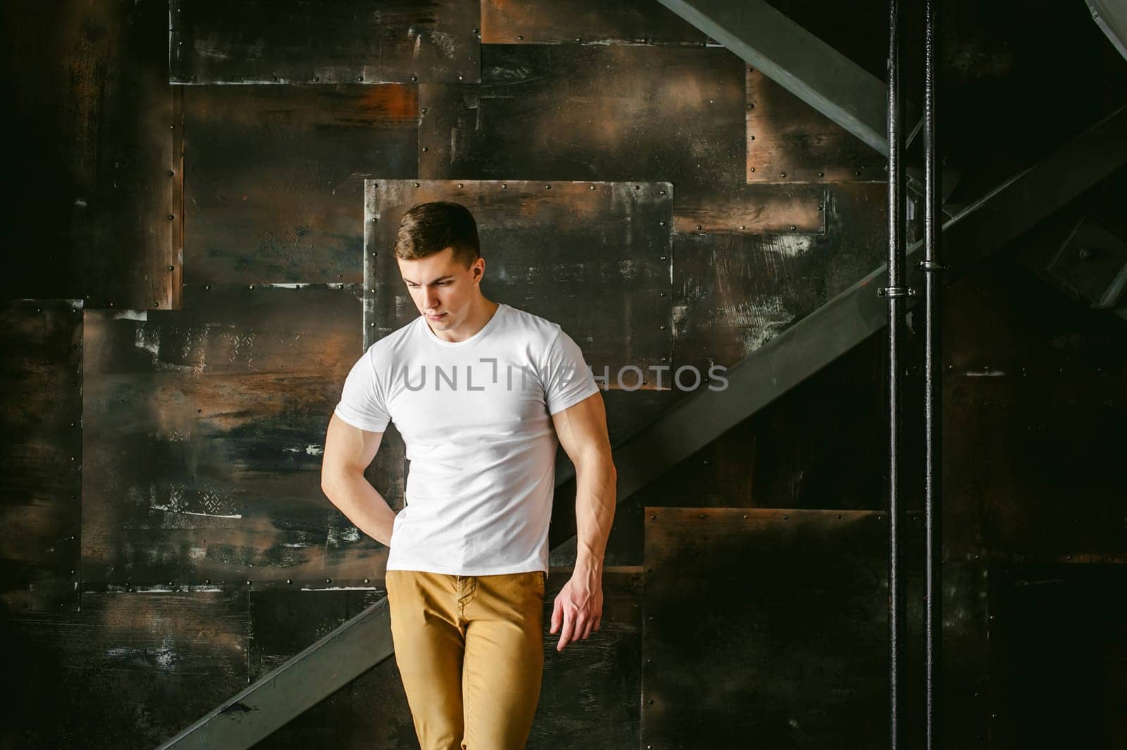 Young sexy men bodybuilder athlete, Studio portrait in a loft on the background of a stylized metal wall, a guy model in a white T-shirt and brown trousers