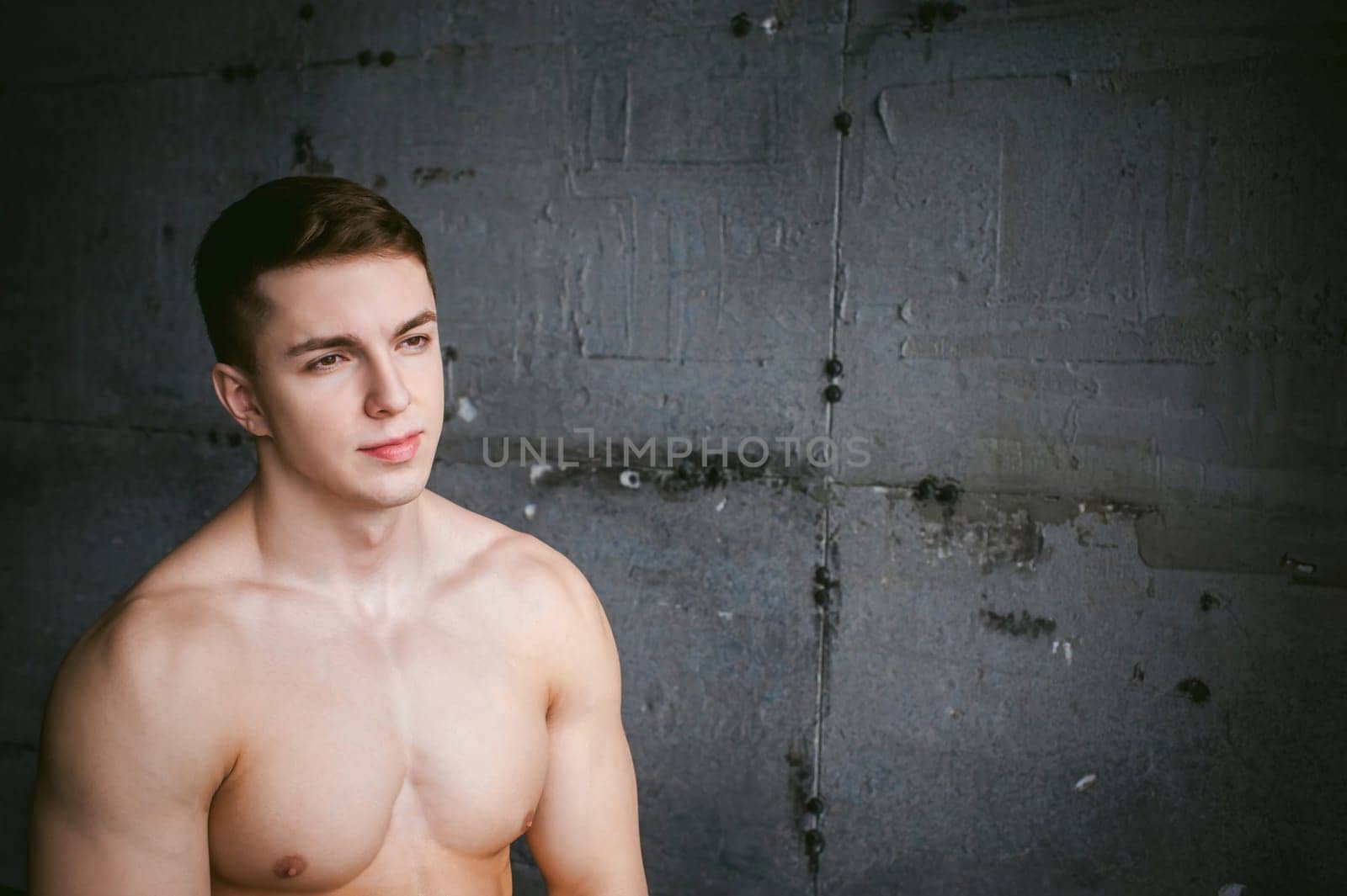 Young sexy male bodybuilder athlete with bare torso against a gray brutal concrete wall, studio portrait of a light-skinned strong man