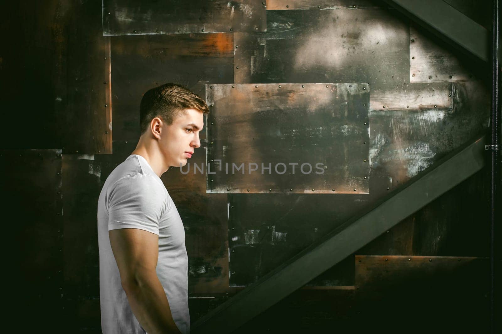 Young sexy men bodybuilder athlete, Studio portrait in a loft on the background of a stylized metal wall, a guy model in a white T-shirt and brown trousers