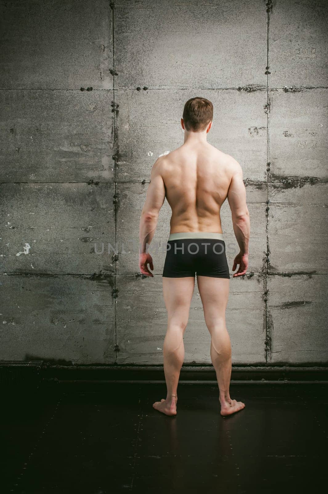 Young sexy male bodybuilder sportsman with a bare torso against a gray wall background, studio portrait of a light-skinned strong man in full growth Stands with his back to the camera