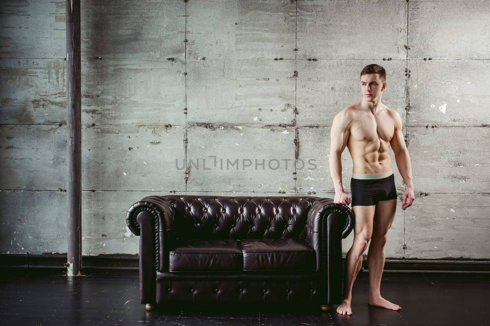 Young sexy men bodybuilder athlete stands at black leather couch with a naked torso, studio portrait