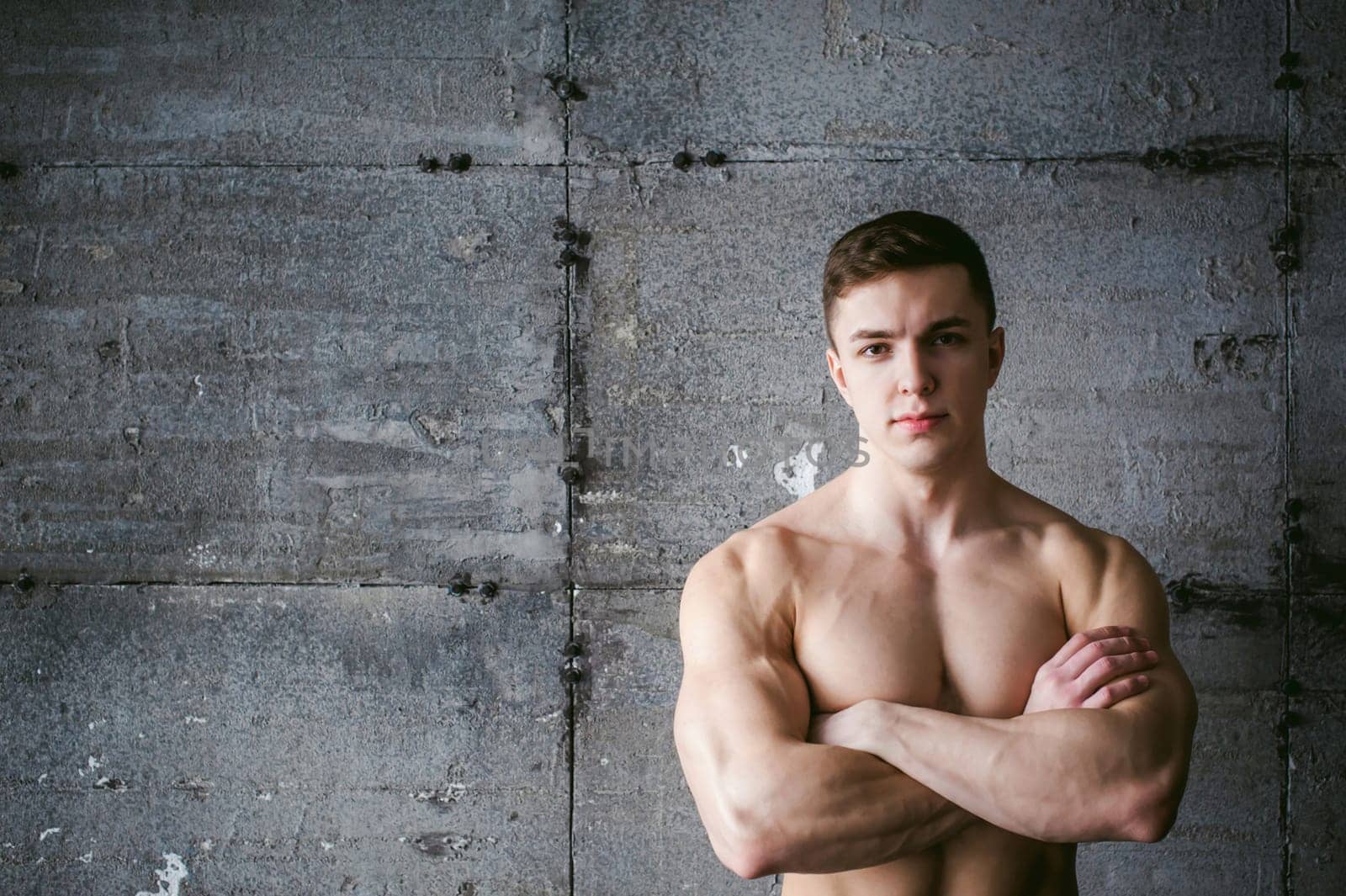 Young sexy male bodybuilder athlete with a bare torso against a gray brutal concrete wall, portrait of a studio of a light-skinned strong man crossed powerful arms on his chest