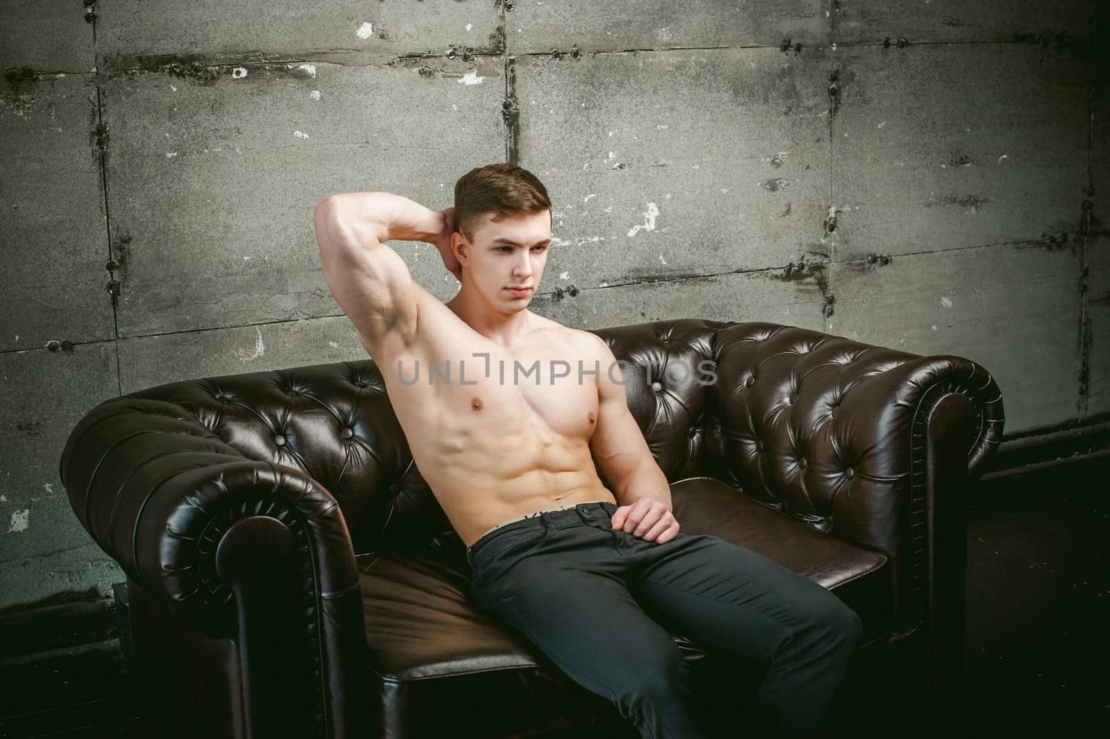 Young sexy men bodybuilder athlete sitting on a black leather couch with a naked torso and black trousers, Threw one's hands behind his head, studio portrait