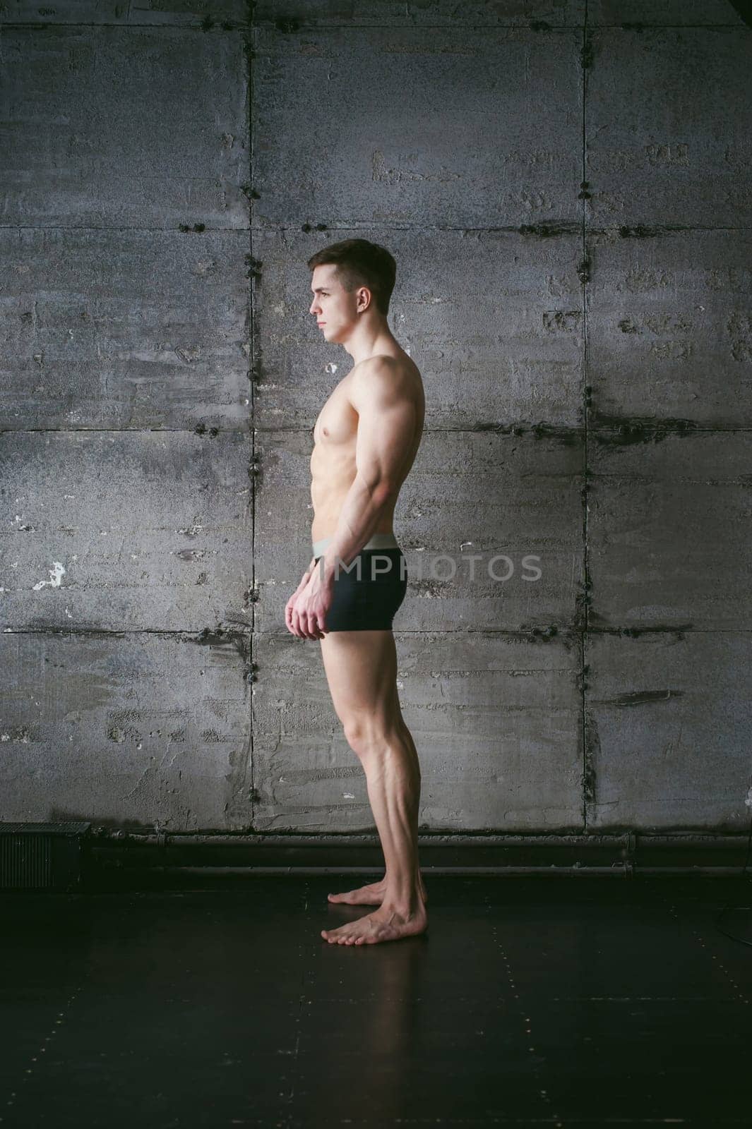 Young sexy male bodybuilder sportsman with a bare torso against a gray wall background, studio portrait of a light-skinned strong man in full growth